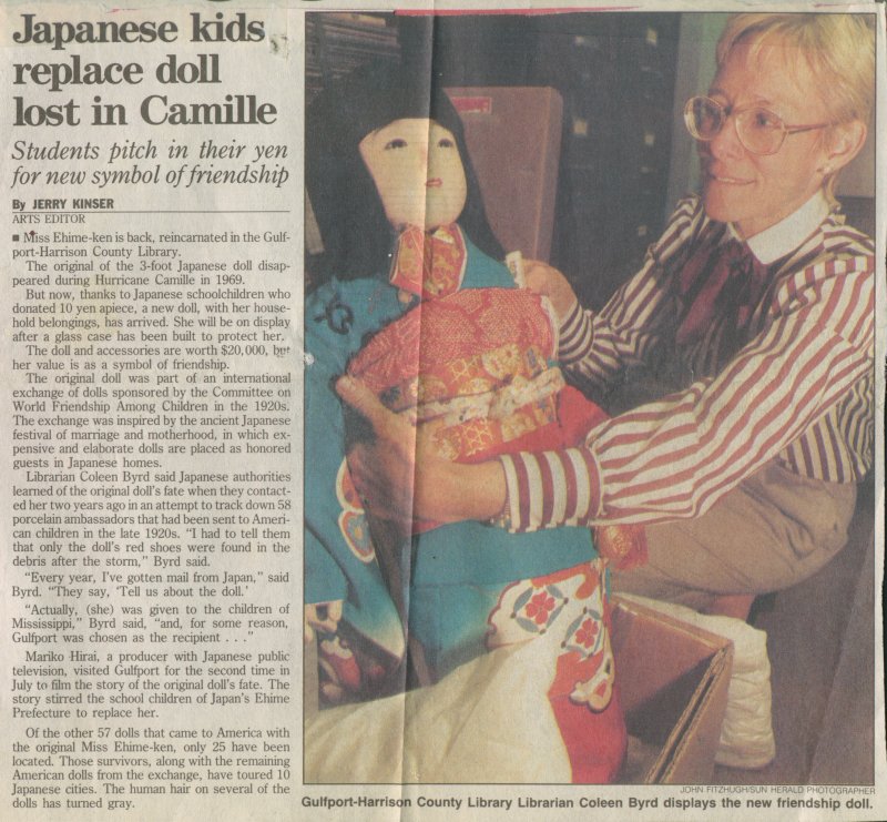 A newspaper clipping titled “Japanese kids replace doll lost in Camille” with the subheading “Students pitch in their yen for new symbol of friendship.” The article describes how Japanese schoolchildren donated a new friendship doll to replace one lost during Hurricane Camille in 1969. The photo shows a person holding a traditional Japanese doll dressed in a colorful kimono, seated in a box.