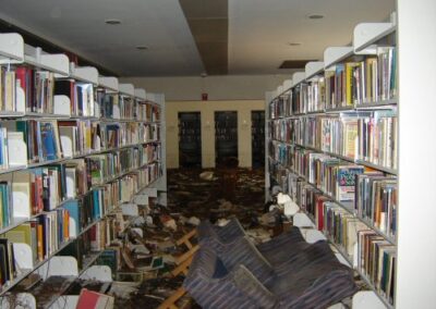 A library aisle lined with tall bookshelves, with the floor covered in mud, soaked books, and overturned furniture. Debris and damaged items are scattered along the corridor, indicating severe flood impact.