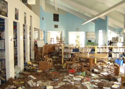 A large library room filled with mud‑covered floors, scattered books, broken furniture, and debris throughout the space. Bookshelves and tables remain standing, but the area shows extensive flooding and structural disruption.