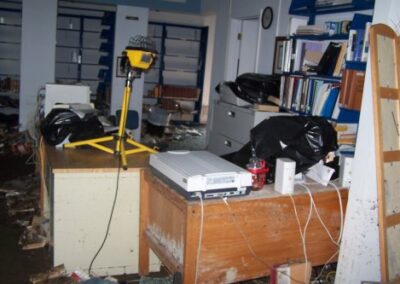 A damaged library office area with desks covered in debris, wires, scattered equipment, and black plastic bags, with muddy floors and items knocked out of place. Shelving units in the background are mostly empty and show signs of water damage.