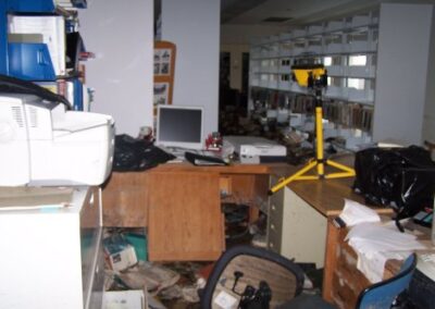 An office area littered with damaged equipment, overturned furniture, and mud‑covered floors, with a work light standing among the debris. Bookshelves and storage areas in the background show signs of flooding.