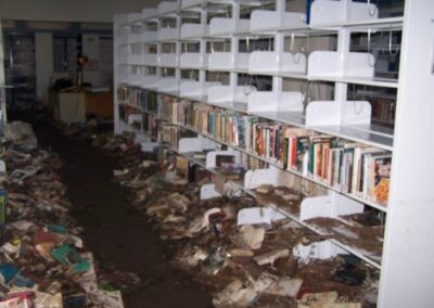 A library aisle filled with mud‑soaked, ruined books scattered across the floor beneath partially emptied shelves. Debris and dirt cover the walkway, showing extensive flood damage.