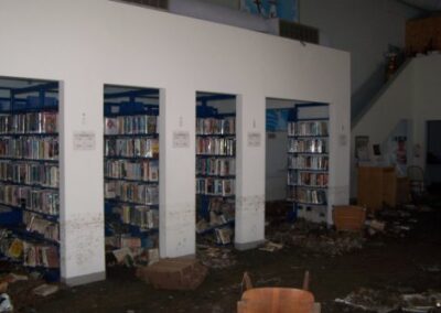 Rows of library shelves stand above a floor coated in mud, fallen books, and debris. Waterlines and dirt stains cover the lower shelves and surrounding walls.