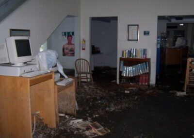 A damaged library area with a computer desk, scattered debris, mud‑covered floors, and furniture knocked out of place. Stains on the walls and cluttered surfaces indicate severe flooding.