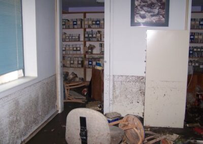 A room with mud‑stained walls and floors shows scattered debris, an overturned chair, and rows of water‑damaged media cases on shelves. A computer desk and cabinet are partially covered in dirt and storm residue.