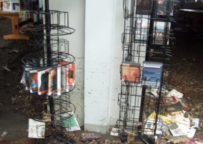 Two metal wire display racks stand in front of a column marked with a high waterline, with mud‑soaked books scattered across the floor. Several books remain on the racks, but most are damaged or displaced by flooding.
