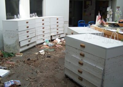 A large room filled with low white cabinets shows heavy flood staining along the drawer fronts and scattered books and debris across the floor. The space appears partially cleared but still shows extensive storm damage.