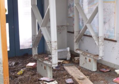 A small entry or seating area filled with mud, splintered wood, and storm debris sits beneath windows marked with a distinct floodline. Furniture pieces are overturned and coated with dirt.