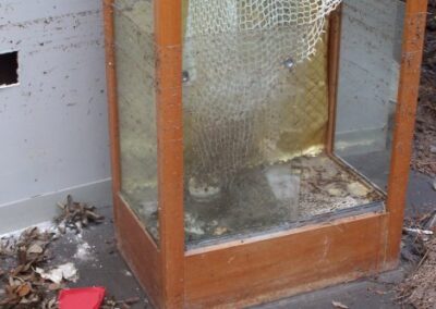 A glass display case with shattered and caved‑in panels stands on a dirty, debris‑strewn floor. The wood frame is water‑damaged, and the interior is filled with mud and broken glass.