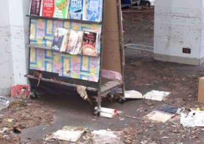 A rolling book display stands near a column stained with mud while water‑damaged books lie scattered across the debris‑covered floor. Leaves and dirt surround the area, indicating heavy flooding.