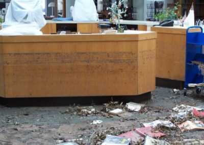 A wooden circulation desk is marked with high floodwater stains, surrounded by piles of damaged books, leaves, and debris on the floor. Items on the counter are covered for protection, indicating post‑storm cleanup.