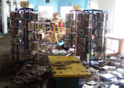 Two rotating book display racks stand in front of heavy storm debris, with mud‑soaked books scattered across the floor and damaged furniture visible in the background. Floodwater stains cover the lower shelving and surrounding area.
