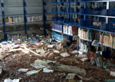 A library room with blue metal bookshelves shows mud‑covered floors, soaked books scattered everywhere, and storm debris pushed into piles. Shelves are partially emptied and stained by floodwater.
