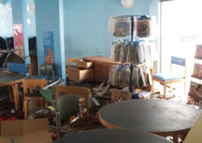 A children’s library area is cluttered with overturned chairs, scattered books, storage bins, and debris, with floodwater stains visible on the walls. Hanging book bags and display racks show signs of storm damage.