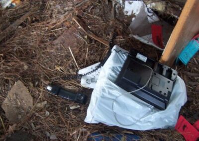 A broken telephone and handset lie in a pile of wet debris and pine straw on the floor. Nearby paper and storm‑damaged items are scattered around the area.