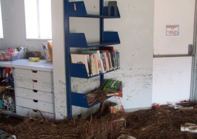 A children’s area with blue metal wall shelves shows heavy flood staining, with pine straw, mud, and soaked books piled across the floor. Cabinets and drawers nearby are filled with water‑damaged items.