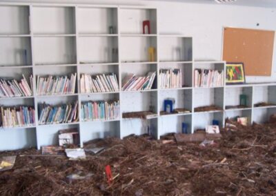 A row of white shelving units filled with children’s books stands above a thick layer of pine straw, mud, and storm debris covering the floor. Several books are warped or scattered, showing widespread water damage.