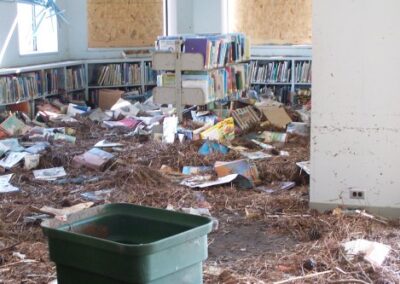 A children’s library room with boarded windows is filled with piles of soaked books, pine straw, and mud covering the floor. Low shelves around the room are stained by floodwater and missing many books.