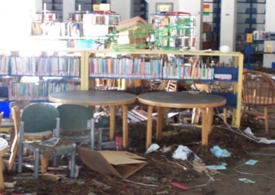 A children’s reading area filled with round tables and chairs is surrounded by scattered books, debris, and mud‑covered floors. Shelving units in the background show stained lower shelves from flooding.