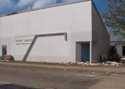 The exterior of the Biloxi Library shows boarded‑up windows, scattered debris along the foundation, and a damaged overhang pipe leaning against the wall. Roofing material appears torn away, and debris lines the sidewalk.