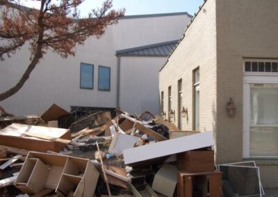 Broken furniture, shelving, and building materials are piled high against the side of the library and an adjacent structure. A damaged tree stands nearby, showing signs of saltwater exposure.