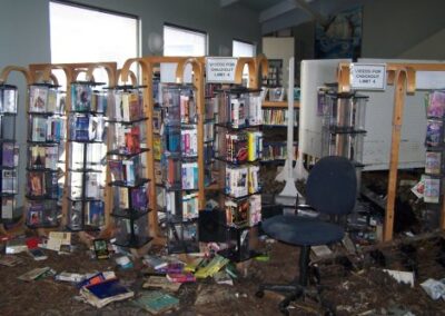 Multiple wooden video display racks stand in a flooded library area, surrounded by soaked books, debris, and a damaged office chair. Mud and storm residue cover the floor and lower shelving.