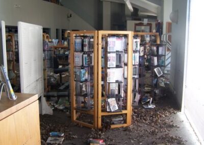 Wooden display racks inside the library hold damaged media items, with leaves, mud, and debris thick across the floor. Additional damaged shelving and furnishings are visible deeper inside the room.