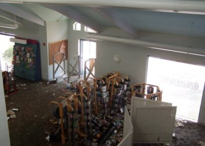 An upper-level view shows a library entry area with damaged media racks, scattered debris, and mud-covered flooring. Bright sunlight enters through a door missing its glass panel, illuminating the destruction.