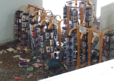 A cluster of wooden media display racks stands in a flooded, debris-covered library area, with scattered books and leaves spread across the floor. A damaged office chair sits near the racks among the storm debris.