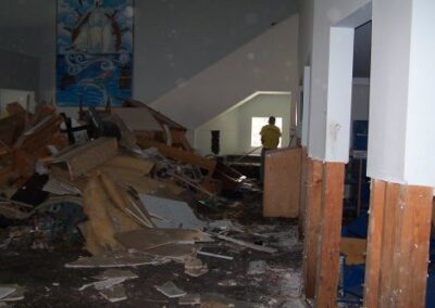 A large pile of broken wood, shelving, and debris fills the center of a damaged library room, while sections of the walls have been stripped to the studs. Mud and storm residue cover the floor throughout the space.
