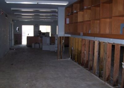 A gutted library room shows lower drywall removed to expose studs, with empty shelving units and debris-stained flooring. Light enters from a distant doorway illuminating scattered equipment.