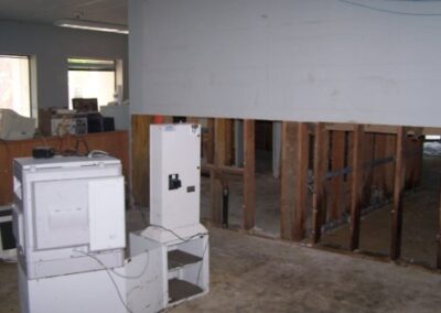 Several pieces of damaged office and computer equipment sit on a bare, debris-stained concrete floor next to walls stripped to exposed studs. Windows in the background show additional storm damage to furnishings.