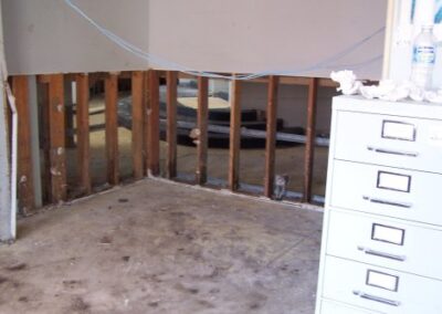 A corner of a damaged room reveals bare studs where drywall has been removed, with debris scattered on the concrete floor and a filing cabinet partially visible in the foreground.