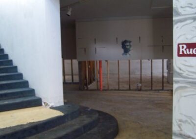 A partially gutted library interior shows exposed wall studs beneath a painted portrait on the far wall, with a staircase to the left and debris‑stained floors throughout.