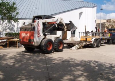 A Bobcat loader is positioned near a flatbed trailer outside the library, surrounded by debris and cleanup equipment. Sunlight and tree shadows fall across the pavement where restoration work is underway.