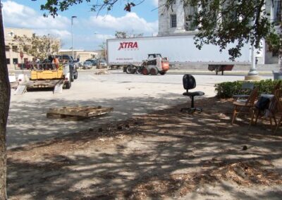 An outdoor area near the library shows scattered debris, a rolling office chair, and construction vehicles operating nearby under sunny skies. Trees cast shadows across the dirt‑covered ground.
