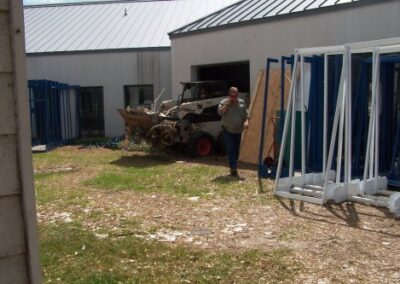A small construction loader is parked near the exterior of the library, with a person walking across the grass toward the equipment. Metal shelving frames are stacked upright along the wall.
