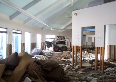 The exterior of a brick building shows a bent handrail, dead shrubs, and a damaged roof structure above. The surrounding area is littered with debris from storm destruction.