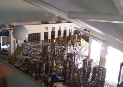 A flooded and gutted library room where drywall has been removed to the studs and debris covers the floor. A utility vehicle sits near the back wall amid the cleanup efforts.