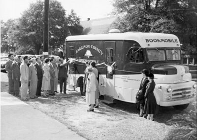 A black‑and‑white photo showing a group of people gathered beside a bookmobile, with a librarian standing at the vehicle's open door as others wait in line.