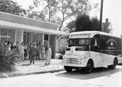 A black‑and‑white photograph showing a bookmobile parked in front of a building while a crowd of people stands nearby, some approaching the vehicle.