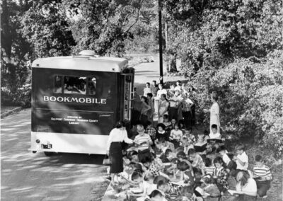 A black‑and‑white photograph showing a bookmobile parked on a tree‑lined street while a large group of children sits on the ground nearby, listening to an adult who appears to be reading or speaking.