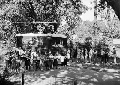 A black‑and‑white photograph of a bookmobile parked along a shaded road as a large group of children gathers around the vehicle and stands in a line nearby.