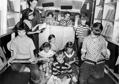A black‑and‑white photograph of children sitting and standing inside a bookmobile, reading books while shelves of books line the walls.