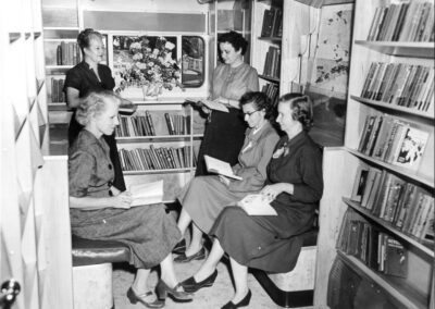 A black‑and‑white photograph of several women seated and standing inside a bookmobile, reading and discussing books while shelves filled with books line the walls.