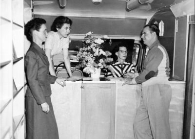 A black‑and‑white photograph of four adults inside a bookmobile, standing and conversing near the front counter with bookshelves lining the walls.