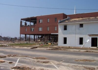 A large brick building on tall stilts shows major structural damage with missing walls and exposed interior spaces. Debris and broken materials are strewn across the adjacent road and lots.