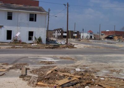 A street lined with damaged buildings and scattered debris shows collapsed structures, broken windows, and piles of wood and rubble across the roadway. Utility poles lean overhead beneath a clear sky.