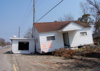A white house sits tilted off its foundation with storm debris scattered around the yard and street. Spray‑painted markings appear on the exterior wall, indicating post‑storm inspection.