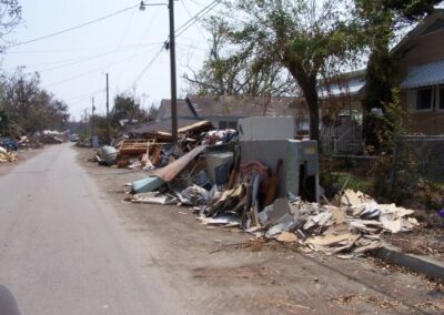 A residential street is filled with storm wreckage, including broken appliances, lumber, and household debris piled along the roadside. Damaged trees and leaning power lines line the quiet neighborhood.